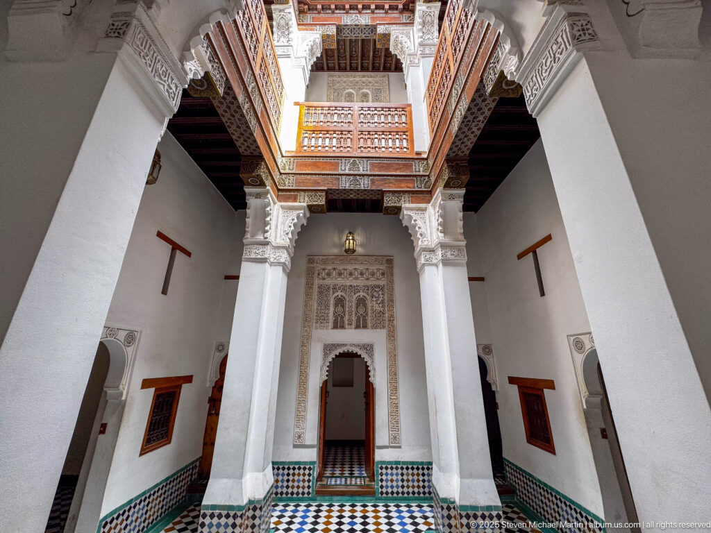 Vertical view of white piers rising from tiled floor and zellij walls to an upper wooden gallery.
