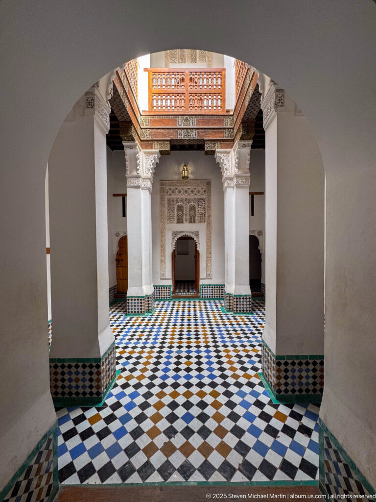 Horseshoe arch frames a tiled interior with geometric zellij pavement, white columns, carved stucco, and a cedar lattice screen.