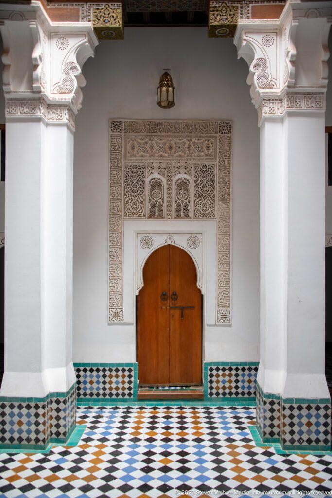 Wooden double door framed by carved white stucco, columns, patterned zellij dado, and a multicolored geometric tile floor.