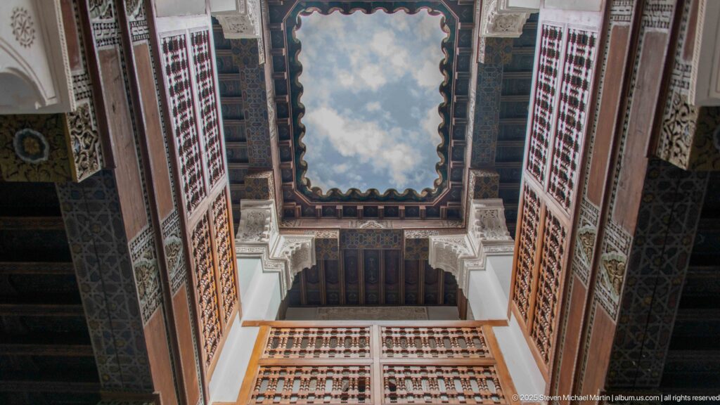 Upward view to blue sky through a rectangular courtyard opening framed by cedar latticework, carved wood, stucco, and balconies.