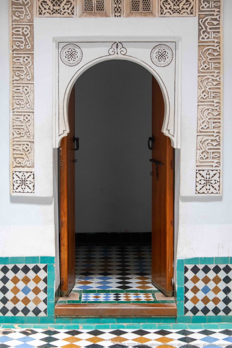 Open horseshoe-arched doorway with wooden doors, carved stucco surround, patterned zellij dado, and tiled threshold.