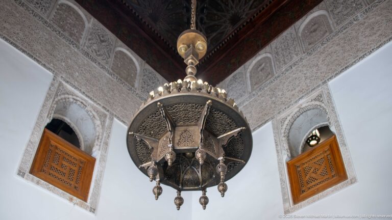 Large pierced-metal hanging lantern beneath dark timber ceiling, carved stucco walls, and small cedar lattice windows.