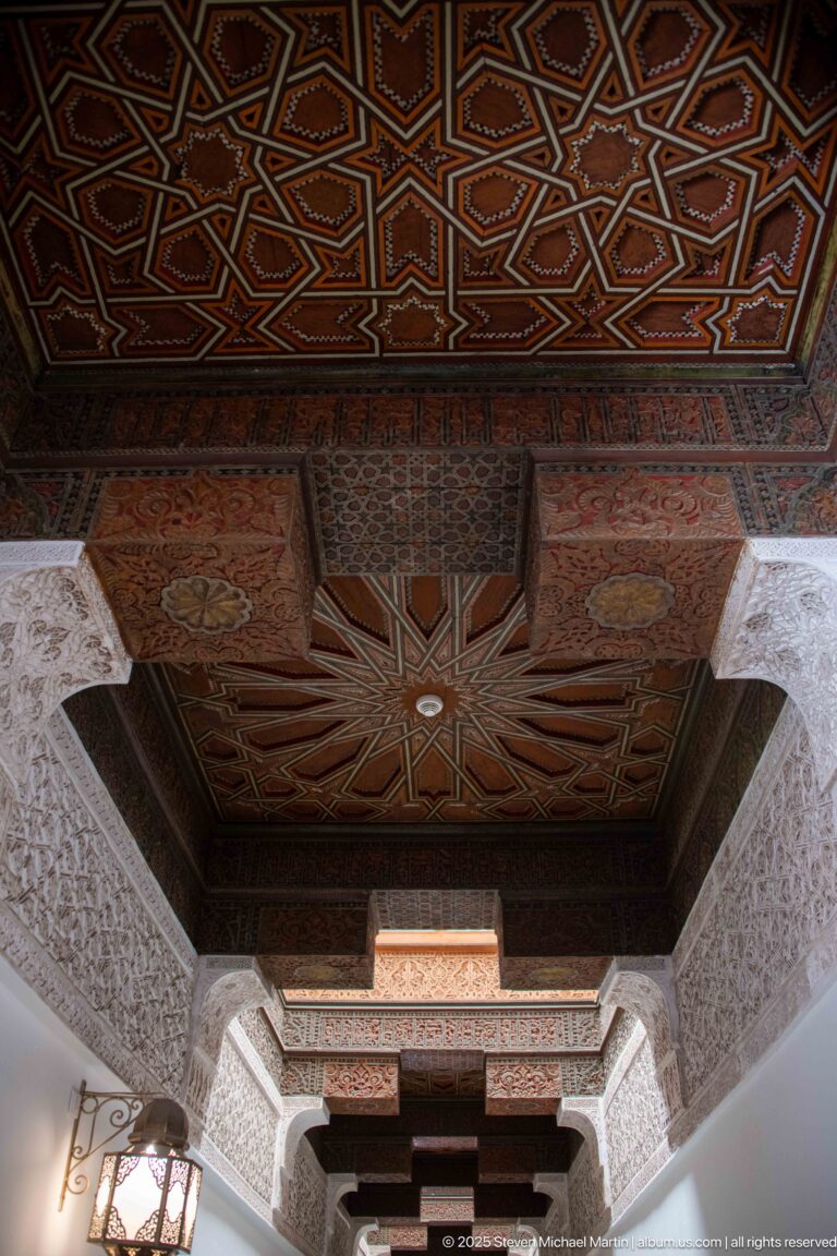 Polychrome wooden ceiling with geometric star panels, carved beams, dark timber, and carved stucco walls along a narrow passage.