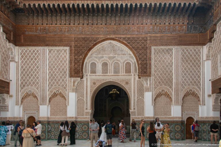 Visitors stand before an ornamented courtyard façade with a tall central arch, carved stucco, woodwork, and zellij panels.