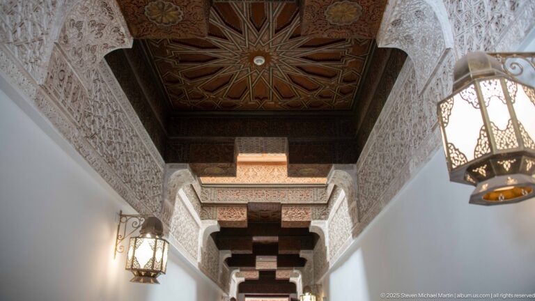 Narrow decorated corridor with carved stucco walls, dark cedar beams, star-patterned ceiling panels, and metal hanging lanterns.