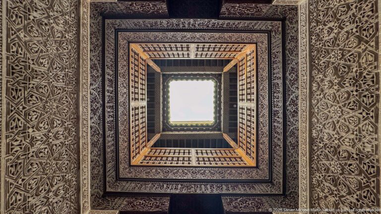 Upward view through concentric square frames of carved stucco, dark wood, and cedar latticework surrounding a bright skylight.