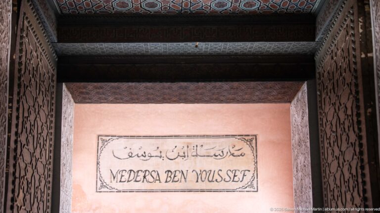 Wall sign reading “MEDERSA BEN YOUSSEF” beneath Arabic script, framed by dark carved wood on a pink wall.