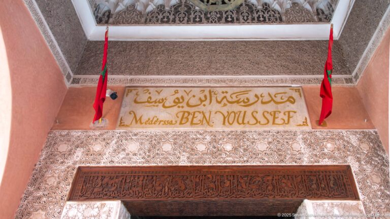 Medersa Ben Youssef entrance sign with Arabic and Latin lettering, two Moroccan flags, carved stucco, and a dark inscription band above the doorway.