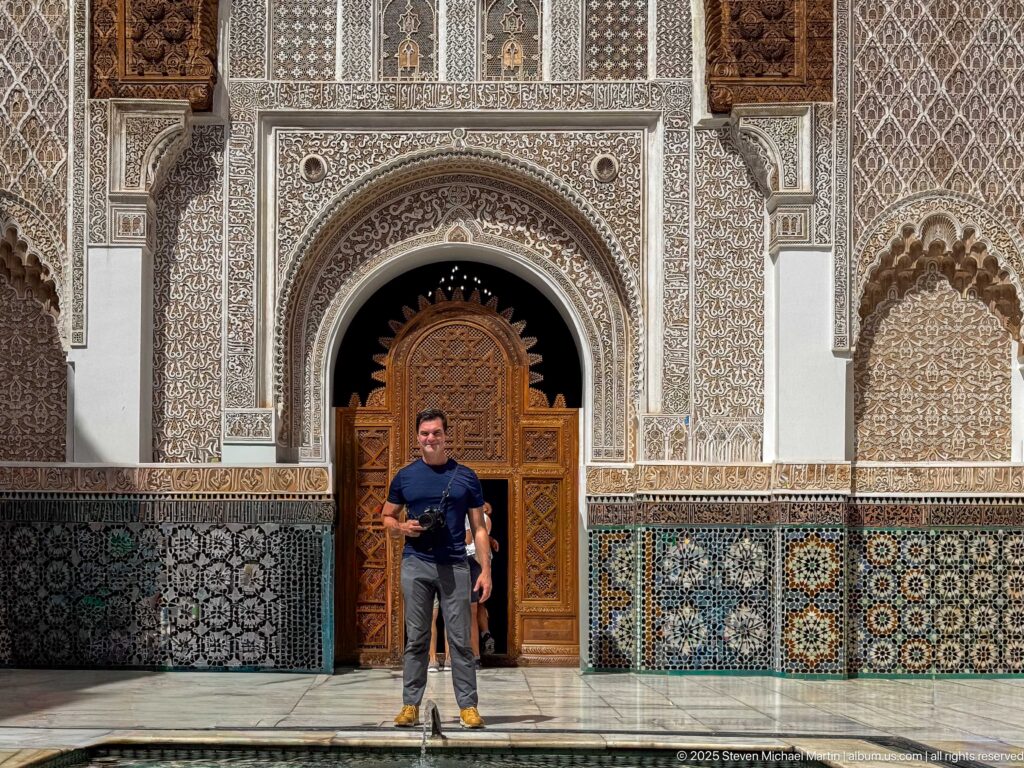 Person standing before a carved wooden doorway framed by ornate stucco and multicolored zellij panels in the courtyard.