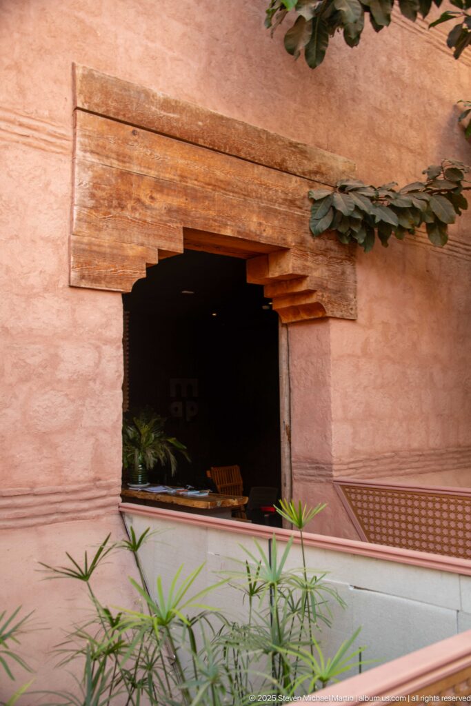Rose-pink plaster courtyard wall with a deep rectangular opening, timber lintels, latticework, and green plants.