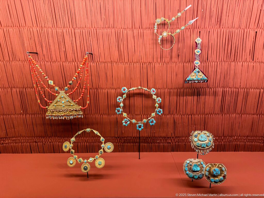 Display of necklaces, earrings, pendants, and round ornaments set with turquoise-blue stones on a terracotta wall.
