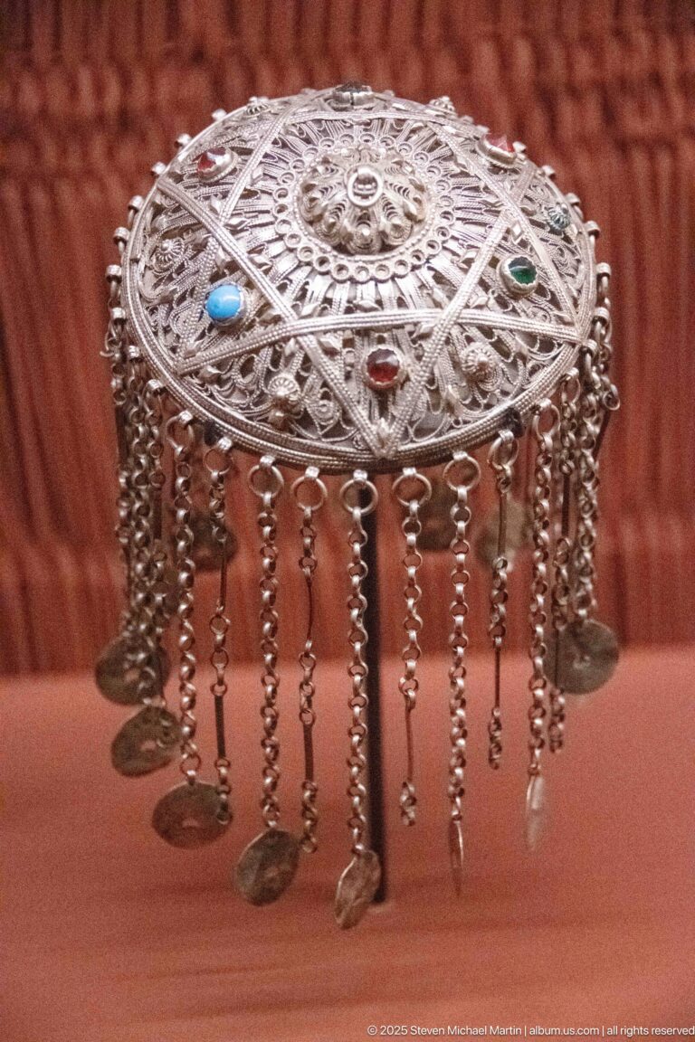 Domed silver-colored ornament with openwork patterns, colored stones, and a fringe of hanging chains and small pendants.