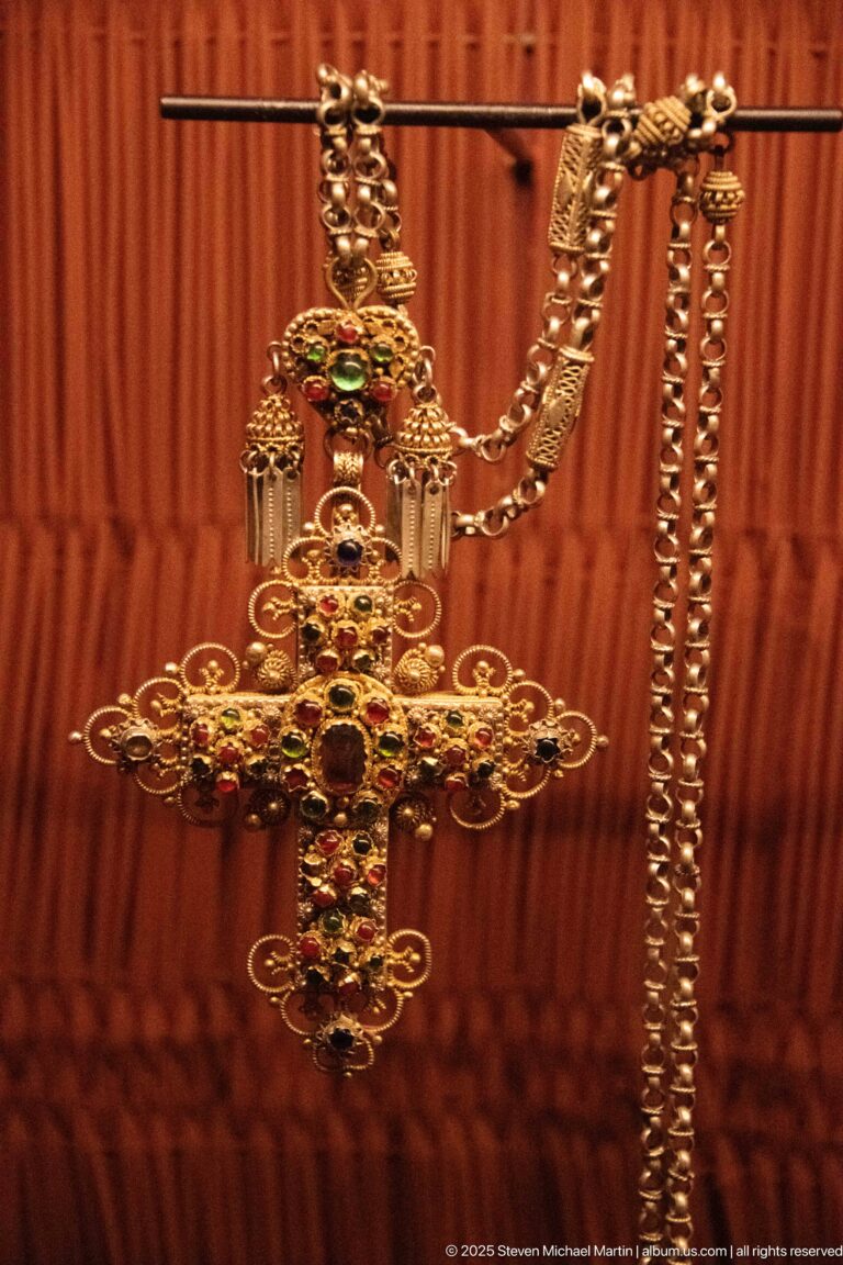 Large ornate cross-shaped metal pendant with red and green inset details hanging from a heavy chain.