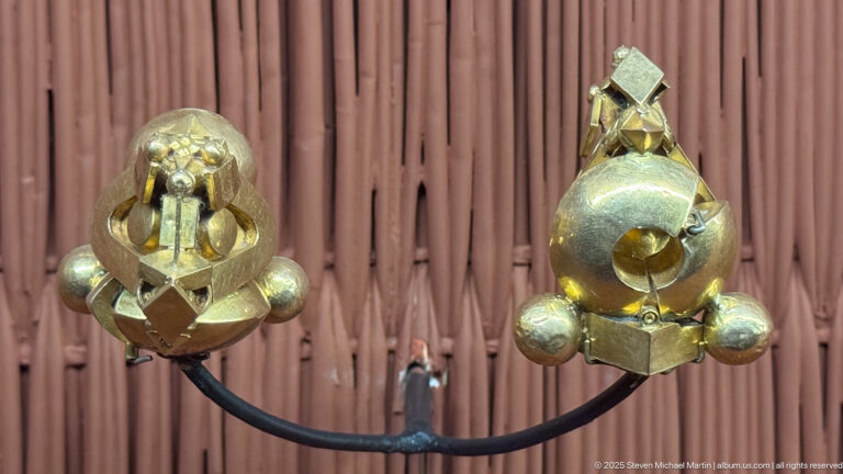 Pair of gold-colored sculptural ornaments with rounded bodies, projecting bosses, and angular upper elements on a curved mount.
