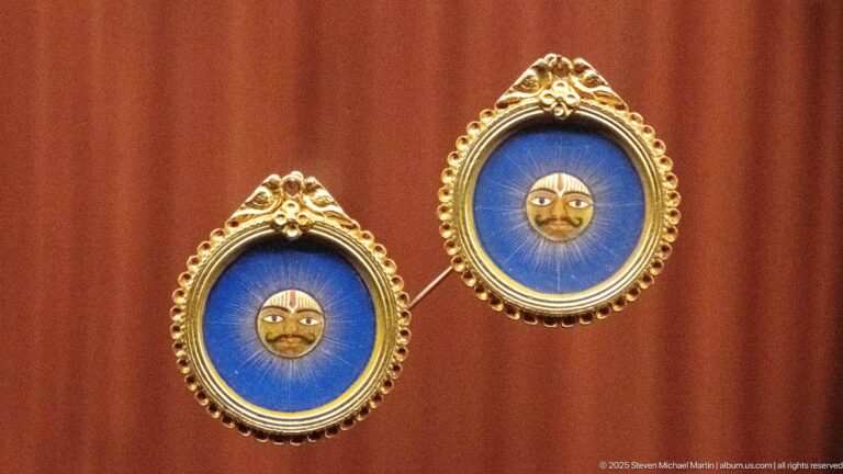 Pair of oval blue earrings with small stylized golden faces, gold-toned borders, and decorative crests.