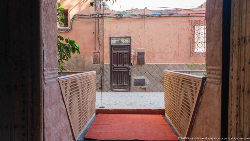 Narrow passage with lattice rails and red runner leading to a dark wooden door in a pink courtyard wall.