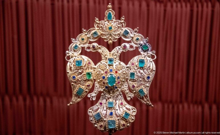 Large openwork metal pendant with symmetrical bird-like forms and blue, green, and red inset stones.