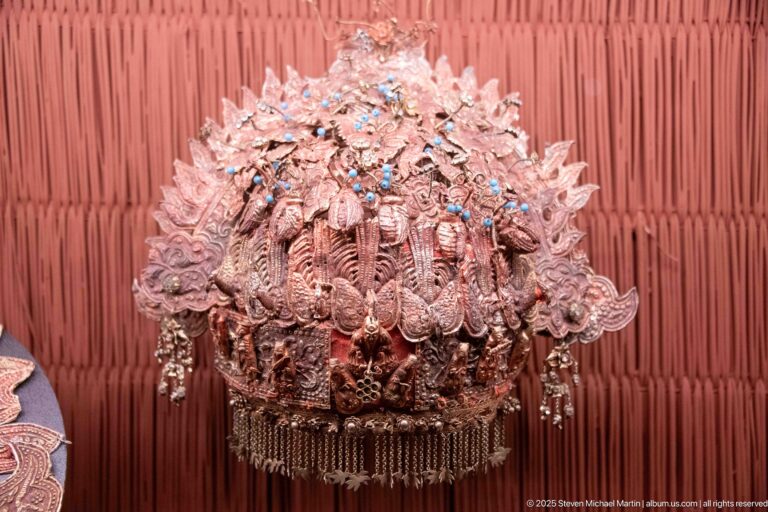 Large domed metal headdress with layered openwork decoration, blue accents, and a dense fringe of hanging pieces.