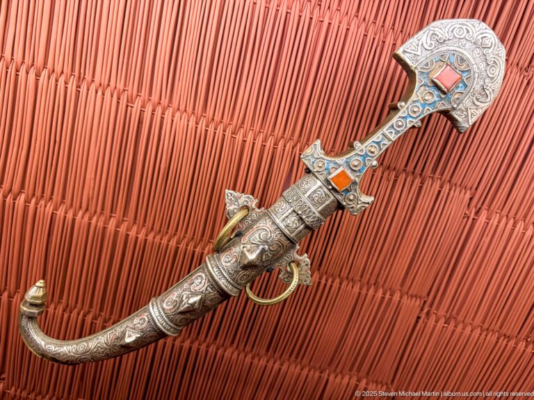 Moroccan curved jambiya-type dagger in an ornate metal-covered sheath with engraved bands, colored insets, and a broad flared hilt.