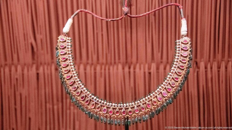 Broad necklace with rows of pink teardrop settings, pale metal borders, and a blue-green hanging fringe.