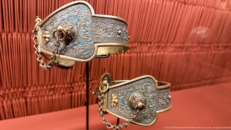 Two broad metal bracelets with blue enamel-like decoration, scrolling patterns, raised bosses, and short chains.