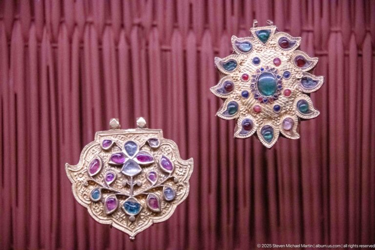 Two ornate metal pendants with engraved surfaces and multicolored stones, one radiating like a flower and one broader and rounded.