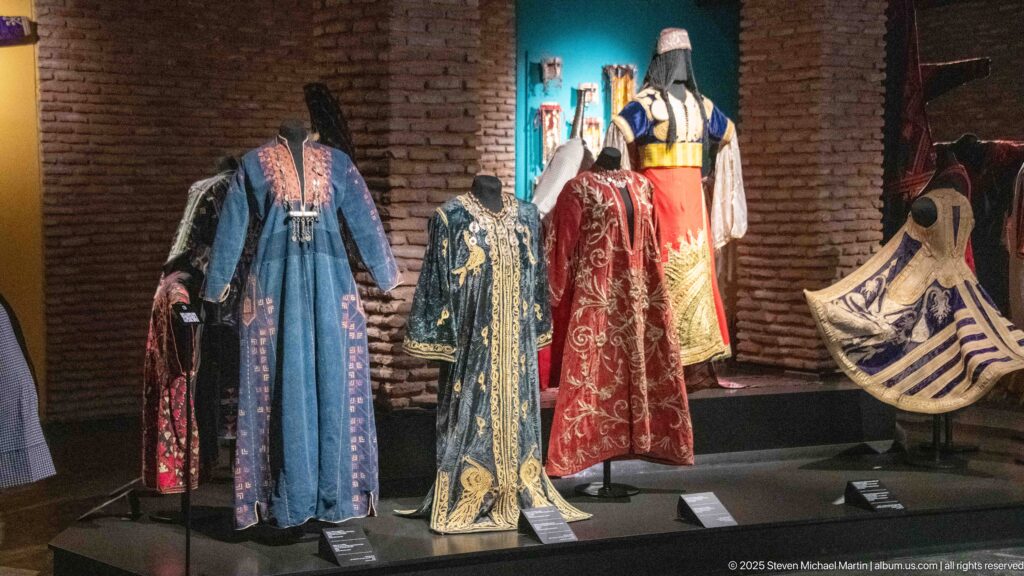 Mannequins wearing long blue, black, red, and cream embroidered robes displayed against brick walls.