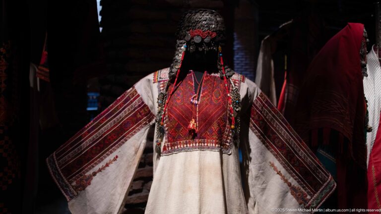 Mannequin in a white robe with red embroidered chest panel, broad decorated sleeves, hanging cords, and an elaborate metallic head covering.