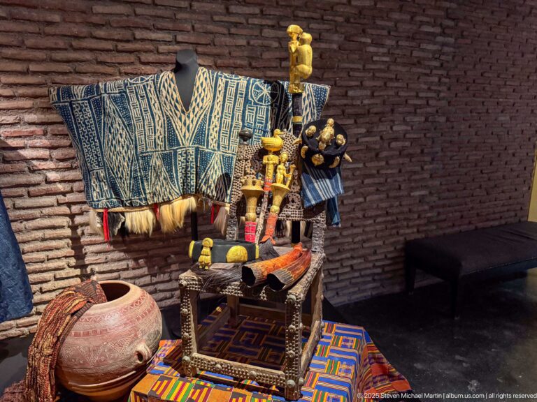 Museum arrangement with blue-and-white woven textile, decorated low seat, pottery vessel, patterned mat, and small objects.