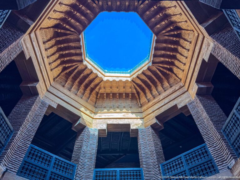 Upward view through MAP Marrakech’s octagonal light well, framed by terracotta brick, timber galleries, lattice screens, and blue sky.