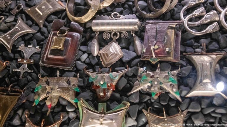 Assorted silver-toned pendants, small boxes, crosses, and geometric amulets arranged on dark pebbles.
