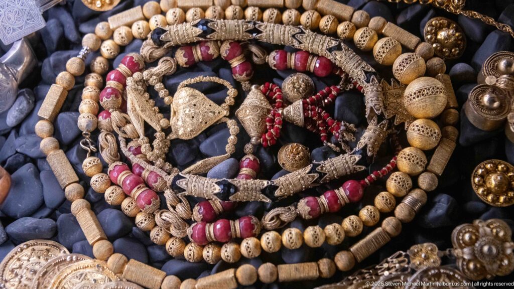 Concentric necklaces of carved cream beads, red beads, braided cords, and metal pendants on dark pebbles.