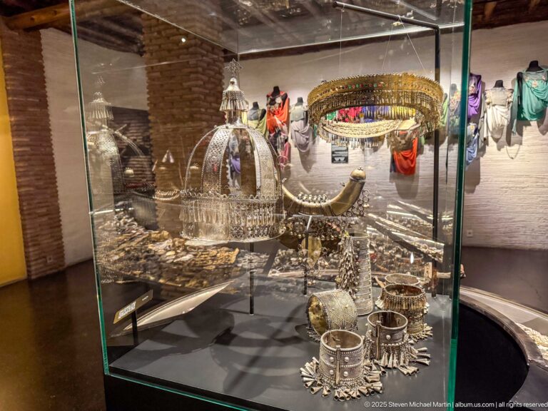 Glass museum case with large domed metal ornaments, bracelets, chains, and smaller jewelry pieces.