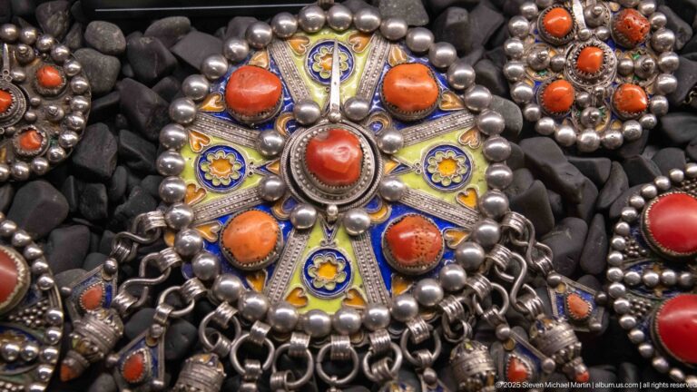 Large circular metal ornament with coral-colored cabochons, yellow-and-blue enamel petals, and chainwork.