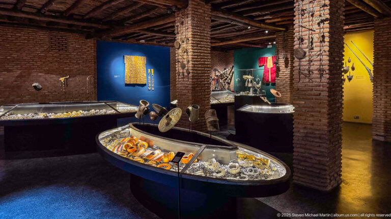 Brick-walled museum gallery with illuminated cases displaying bracelets, necklaces, and metal ornaments.