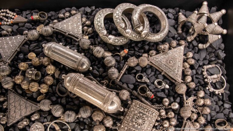 Silver-toned rings, beads, triangular pendants, cylindrical amulet cases, and heavy bracelets on dark pebbles.