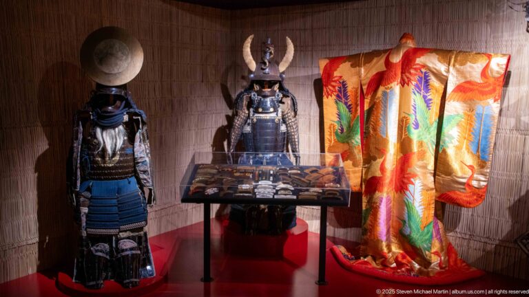 Two Japanese samurai armor ensembles with kabuto helmets, laced lacquered plates, shoulder defenses, and an orange patterned robe beside them.