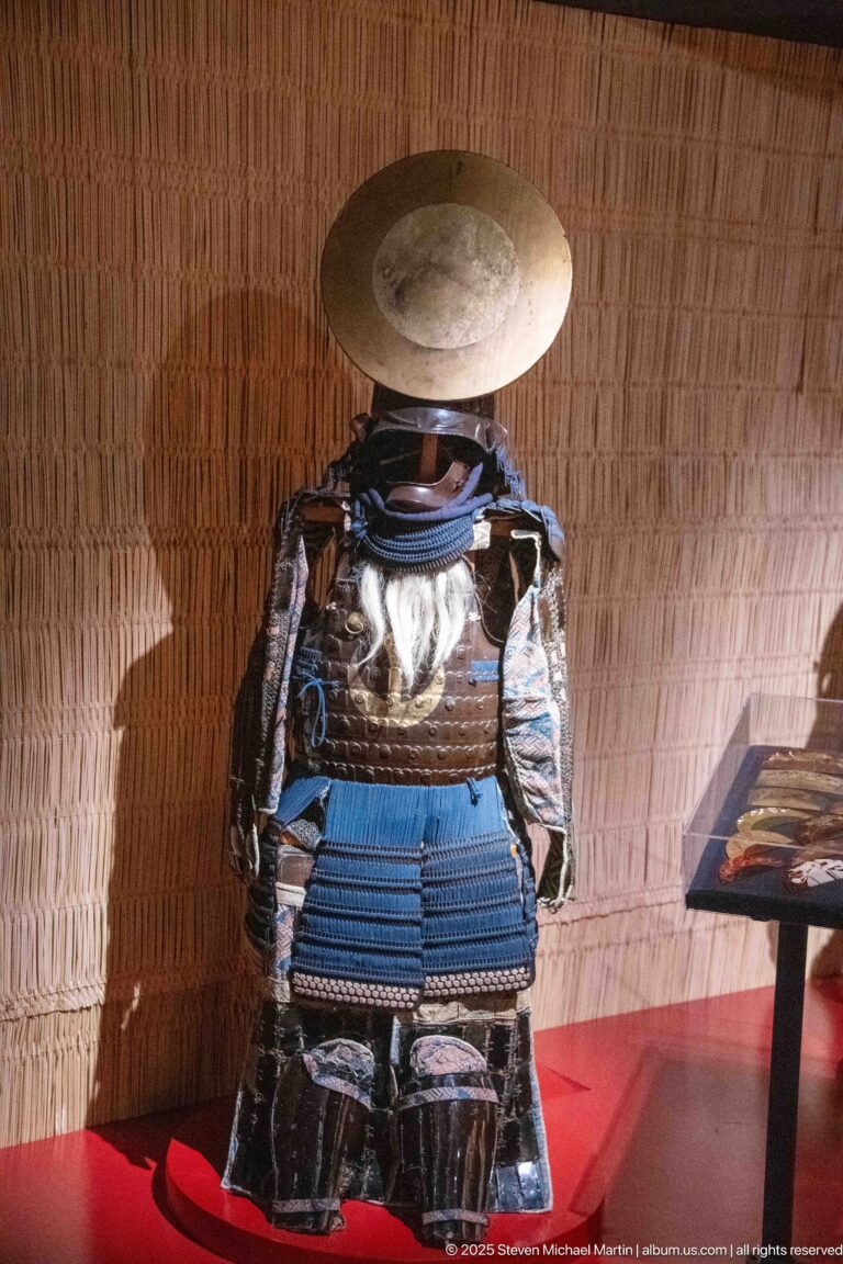 Front view of Japanese samurai armor with blue lacing, dark cuirass, patterned sleeves, and a broad circular helmet crest.