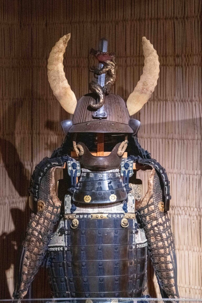 Close view of Japanese samurai armor with a horned kabuto helmet crest, dark cuirass, broad shoulder guards, and patterned sleeves.
