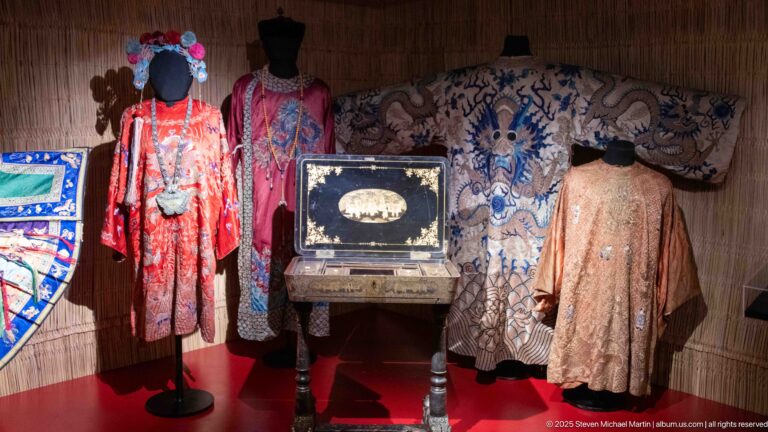 Embroidered East Asian robes beside a pale textile with blue dragon-like motifs and a small decorated chest.