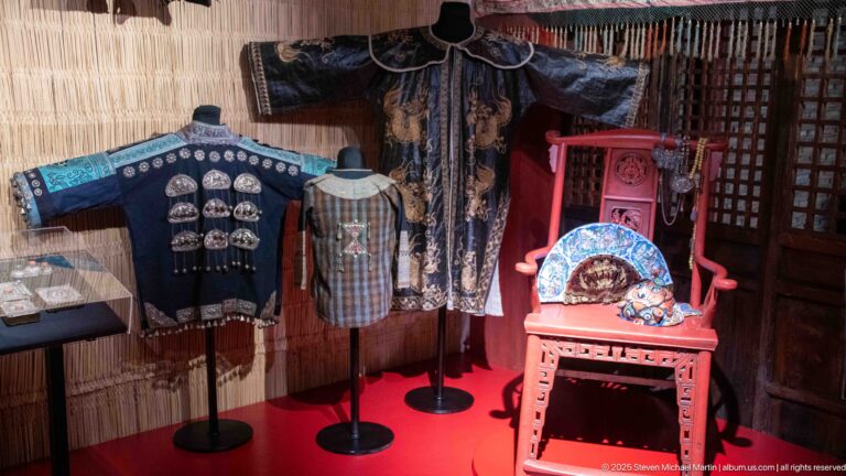 Dark embroidered robes and jackets around a red chair displaying painted folding fans.