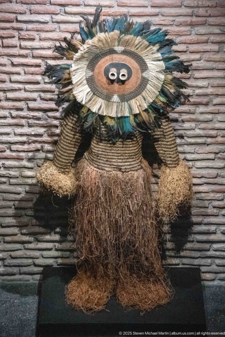 Anthropomorphic display object with woven body, shaggy fiber fringe, and a large circular feathered face or headdress.