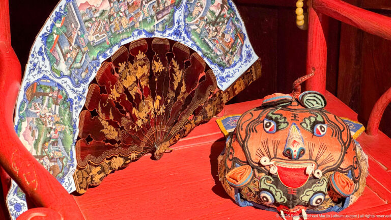 Two decorated folding fans, one with painted scenes and one reddish with gold figures, beside a round painted face object.