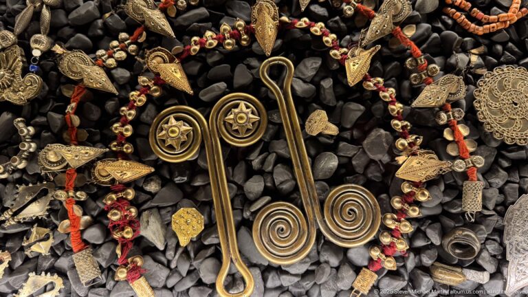 Two large double-spiral metal ornaments inside a necklace of small beads, red accents, and triangular pendants.