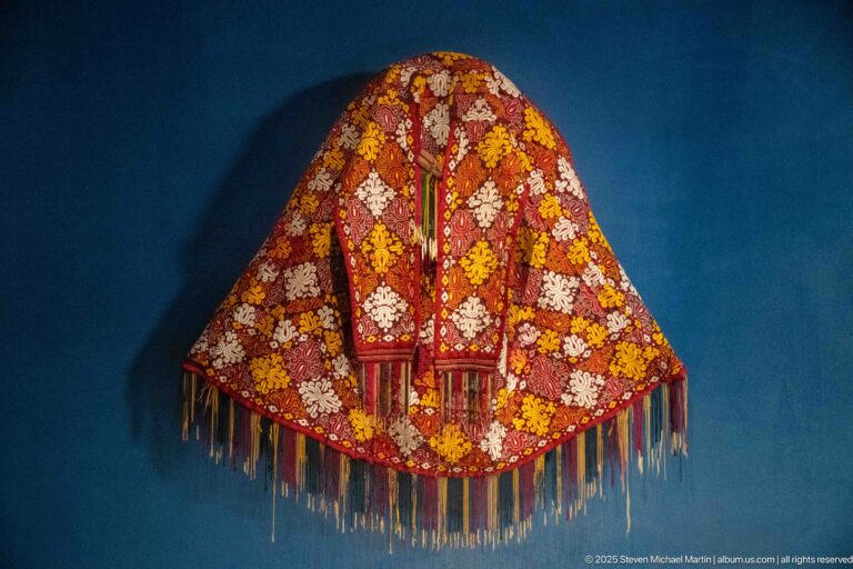 Red-orange embroidered shoulder garment with yellow and white floral motifs, central opening, and multicolored fringe on a blue wall.