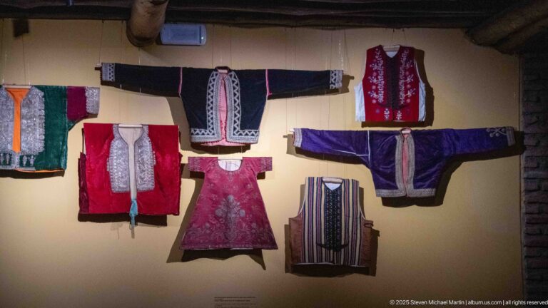 Wall-mounted group of embroidered jackets, tunics, and vests in red, black, purple, green, and striped fabrics.