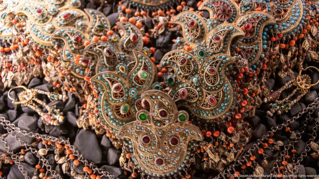 Cluster of paisley-shaped metal ornaments set with turquoise, red, and green stones among orange bead strands.