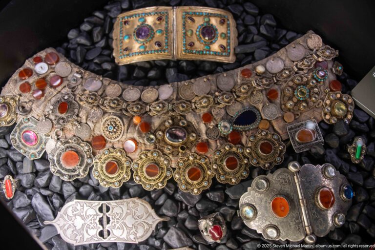 Ornate metal jewelry with multicolored stones, pendants, rings, and linked plaques arranged on dark pebbles.