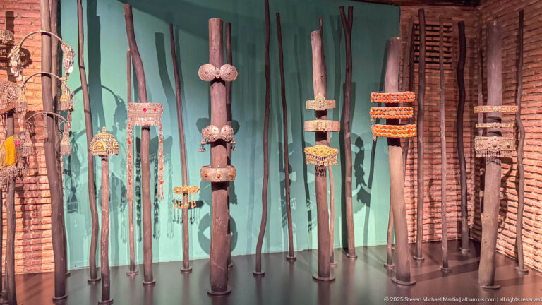 Row of tall decorated wooden staffs or walking sticks with carved, wrapped, and beaded fittings against a turquoise wall.