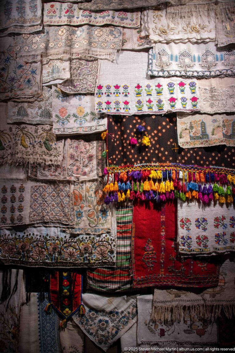 Layered embroidered textiles in red, black, cream, and multicolored patterns with dense fringe and pom-pom tassels.